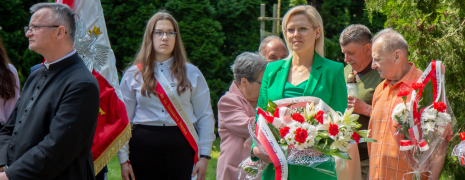 Grupa ludzi stoi na cmentarzu, kobieta trzyma bukiet kwiatów