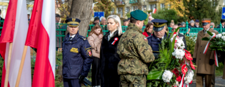 Kobieta stoi w asyście straży miejskiej, przed nimi wojskowy odbiera wieniec
