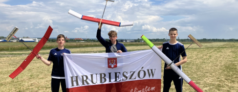 Trzech modelarzy trzyma samoloty a przed nimi flaga hrubieszowa