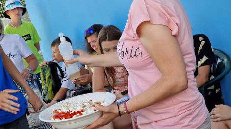 Kobieta trzyma w dłoni talerz z posiłkiem, tłumaczy coś siedzącym obok dzieciom