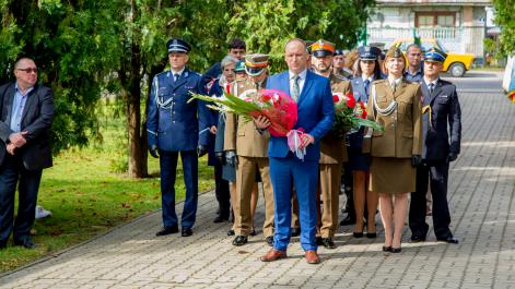 Mężczyzna trzyma wiązankę kwiatów, za nim stoją przedstawiciele jednostek mundurowych