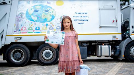 Dziewczynka trzyma w ręku dyplom, pozuje do zdjęcia na tle śmieciarki Beti