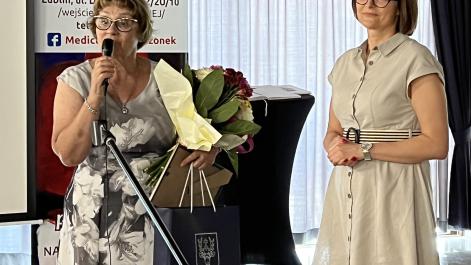 Przewodnicząca stowarzyszenia przemawia przez mikrofon, trzyma w rękach upominek i kwiaty, obok stoi Monika Podolak sekretarz miasta